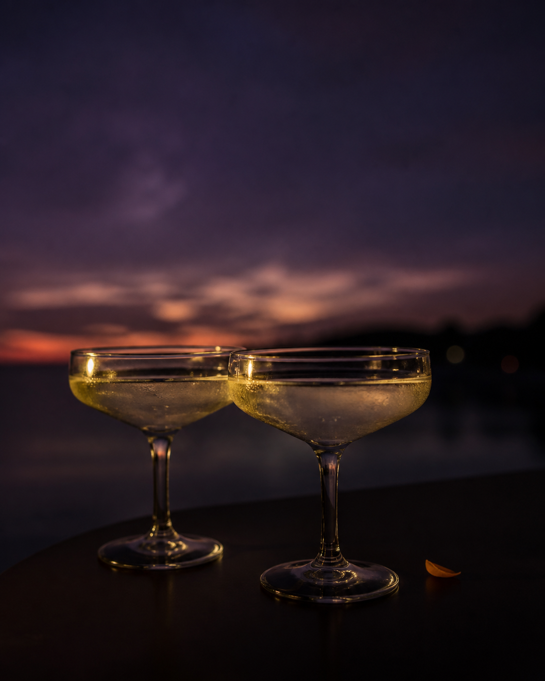 IP-GFT-004 · The celebration — two glasses just poured on a dark table at the blue-gold edge of dusk, warm light catching the rims.