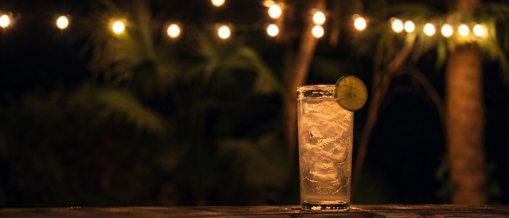 A tall cocktail glowing on a dark porch rail under a haze of warm string-light bokeh.