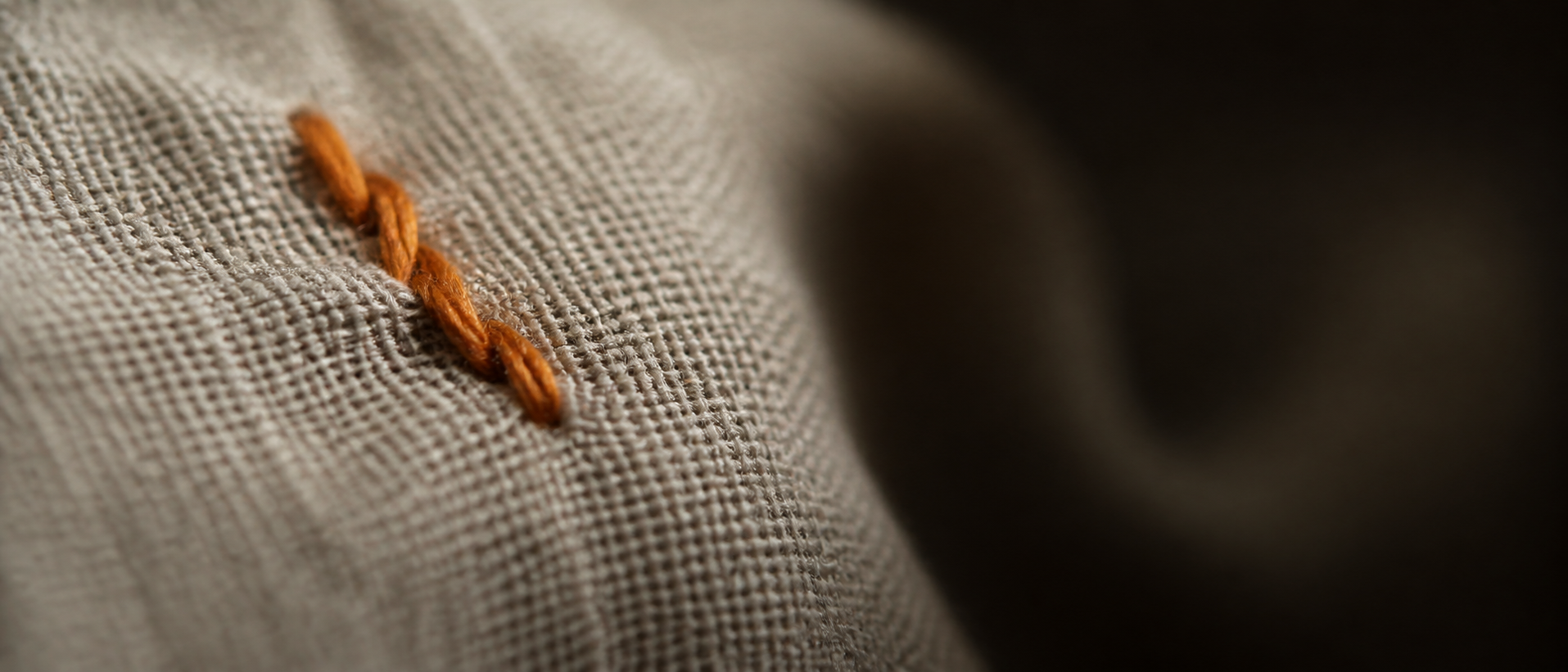 A single marigold-orange thread wound over linen, lit by one warm light against deep shadow.